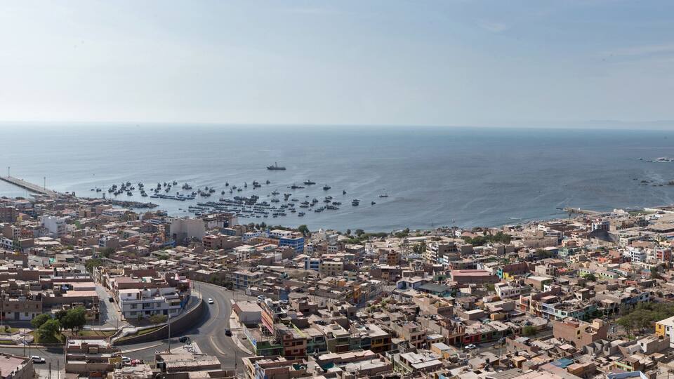A coastal cityscape with dense urban buildings, a harbor filled with boats, and a distant mountain range.