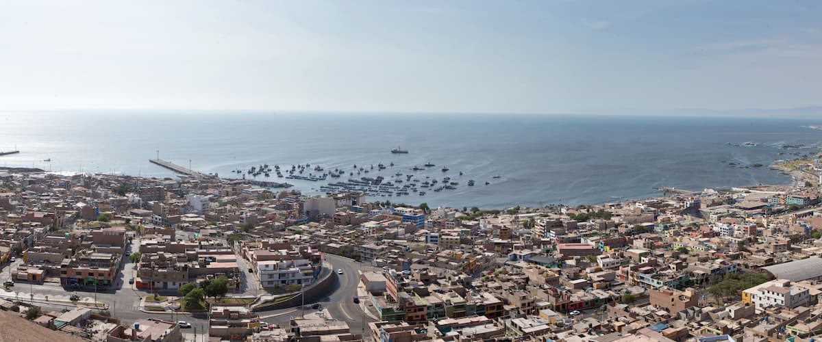 A coastal cityscape with dense urban buildings, a harbor filled with boats, and a distant mountain range.