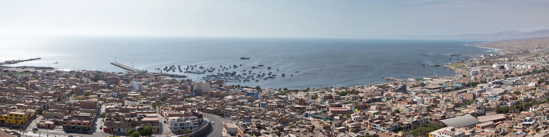 A coastal cityscape with dense urban buildings, a harbor filled with boats, and a distant mountain range.