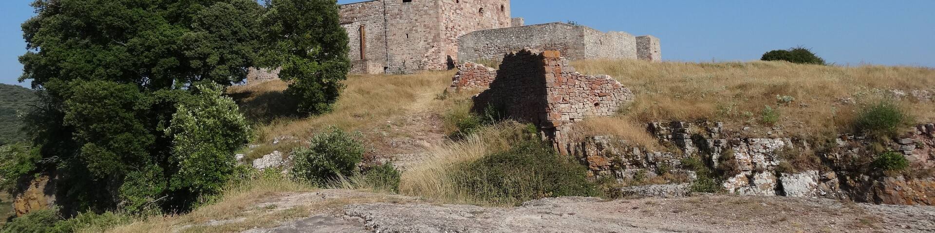 An ancient stone building with a bell tower, surrounded by dry grass and trees on a hill.