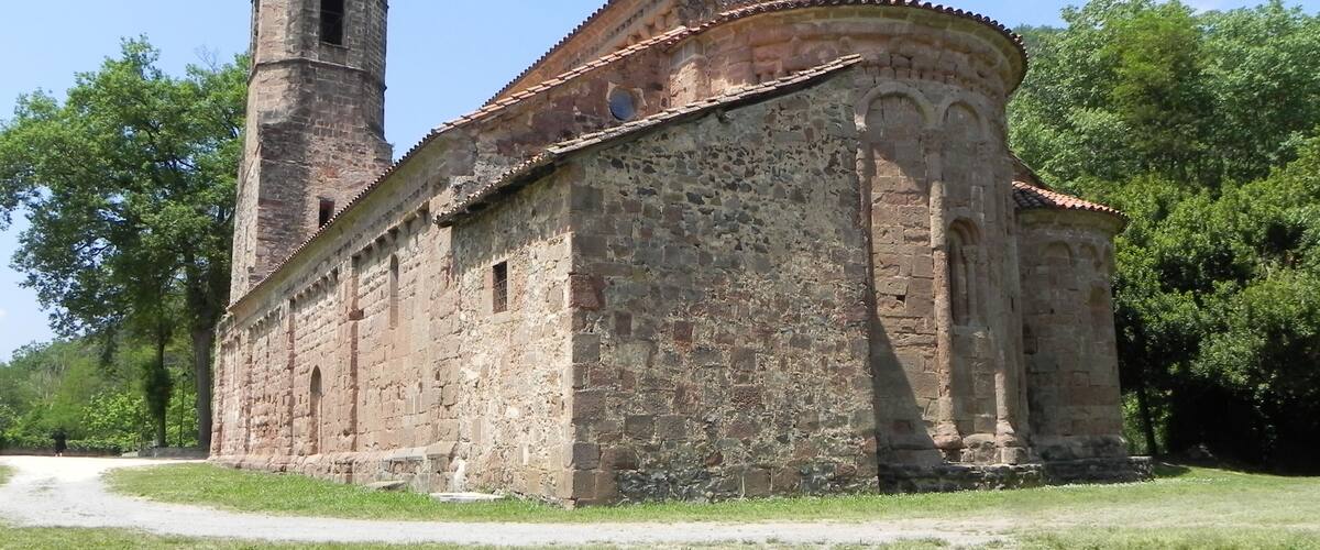 Uma igreja de pedra histórica com uma torre de sino e telhado de azulejos, cercada por vegetação exuberante.