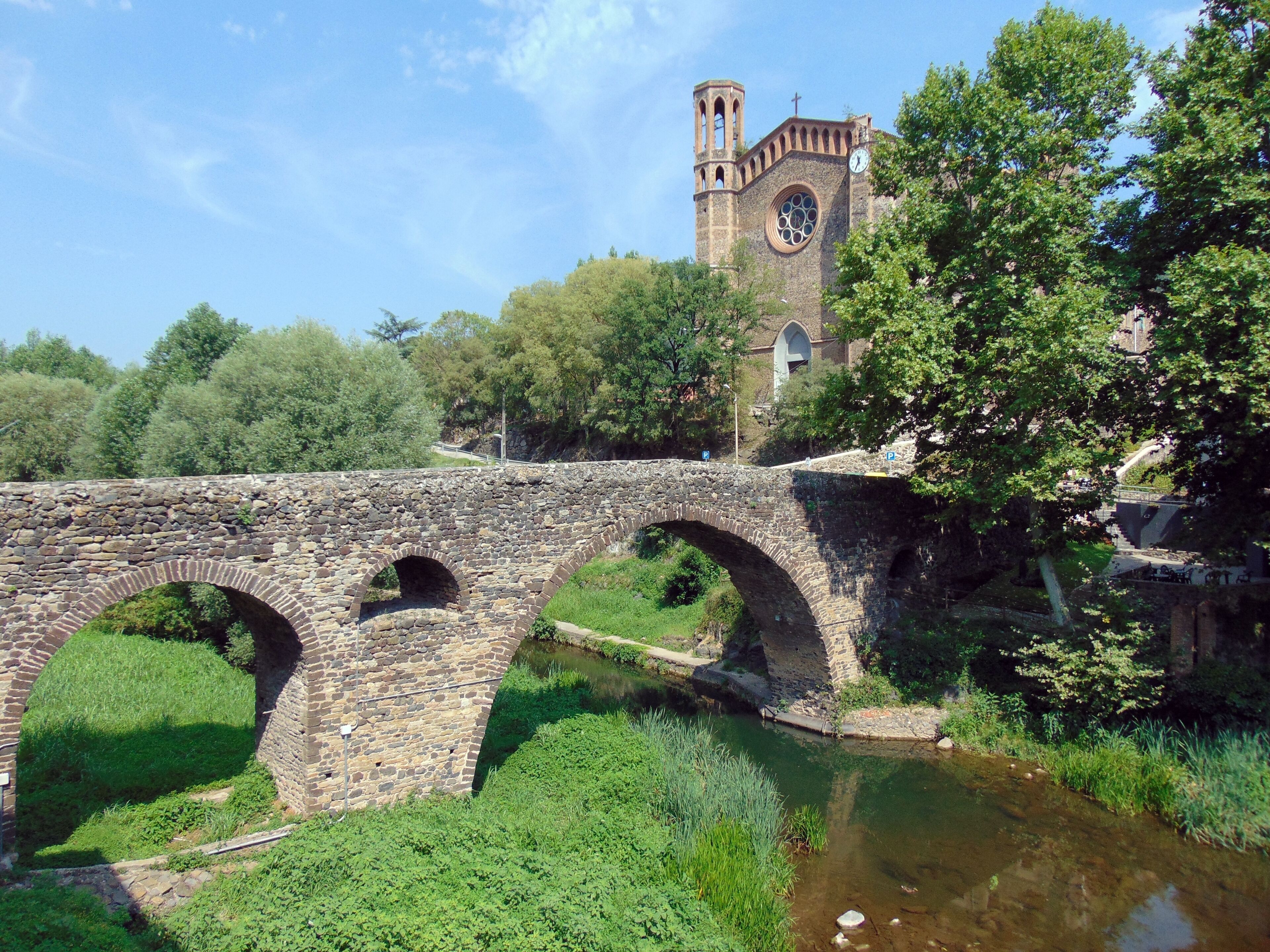 A stone bridge with arches over a river, surrounded by greenery and a church with a clock tower in the background.