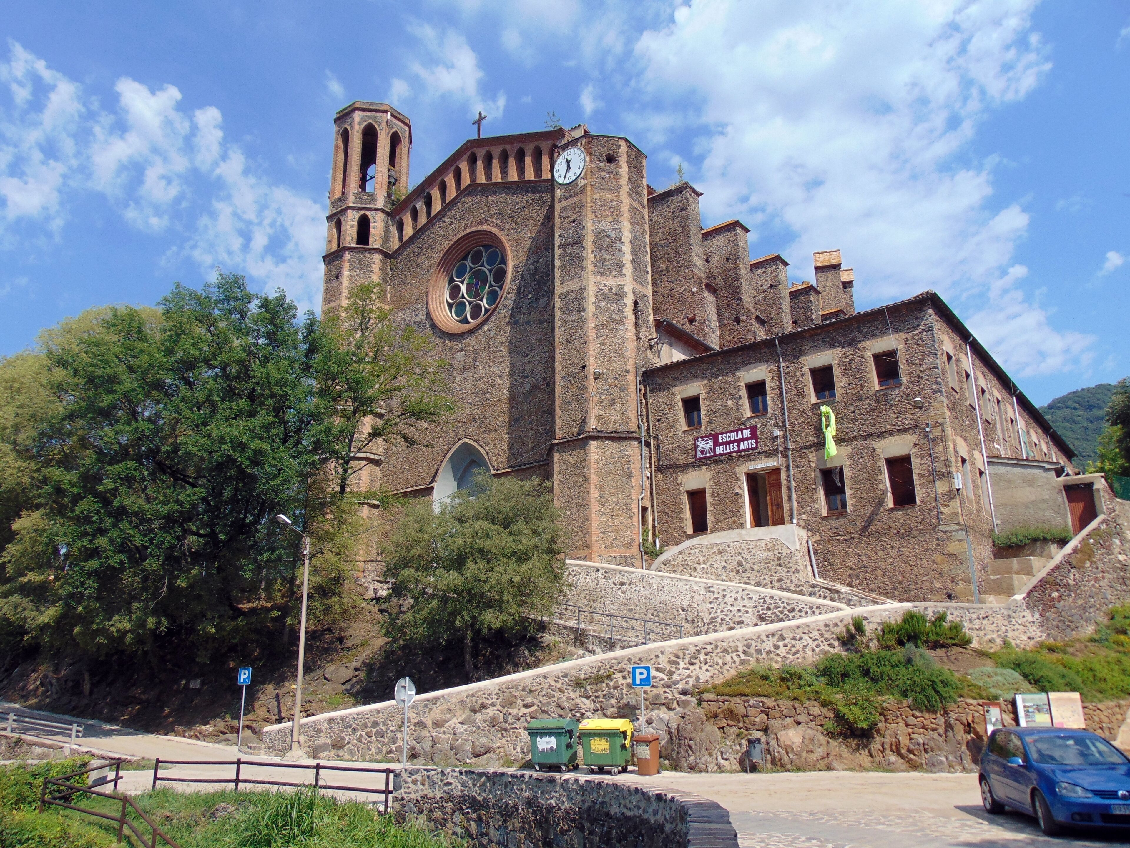 A historic church with a bell tower and a clock, surrounded by trees and a stone wall.