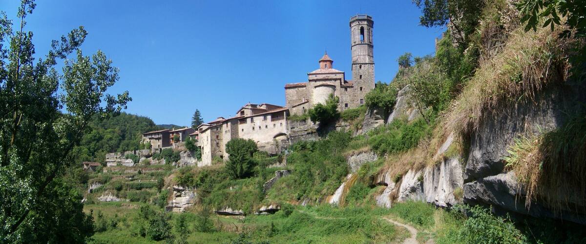 A historic stone church with a bell tower on a hillside, surrounded by lush greenery and a river.