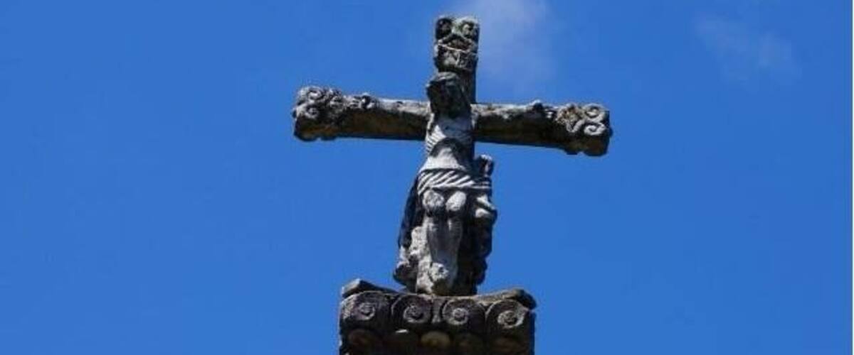 A stone cross with a figure of Jesus Christ, set against a clear blue sky.
