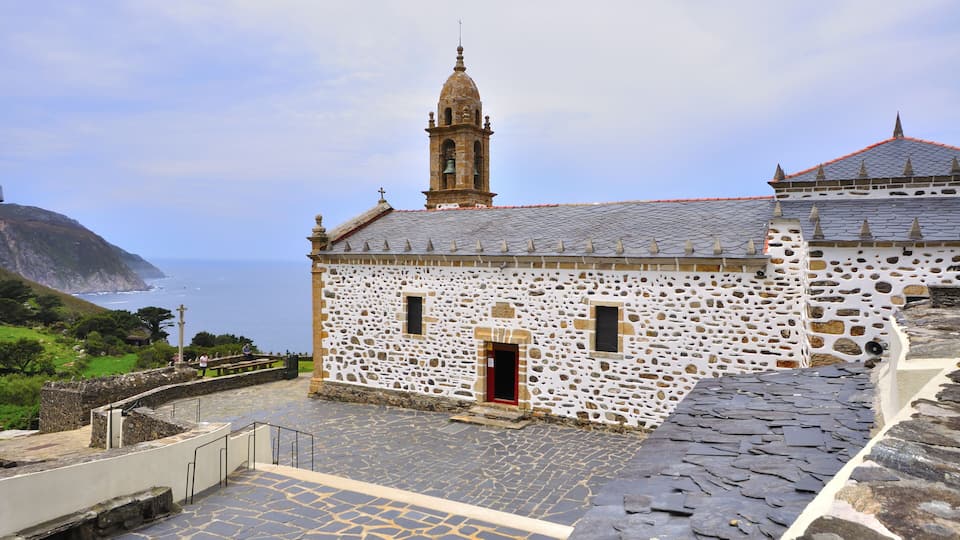 A white stone church with a bell tower and a red-roofed building.