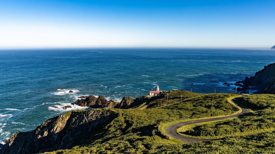 A coastal landscape with a lighthouse on a rocky cliff, a winding road, and the ocean.