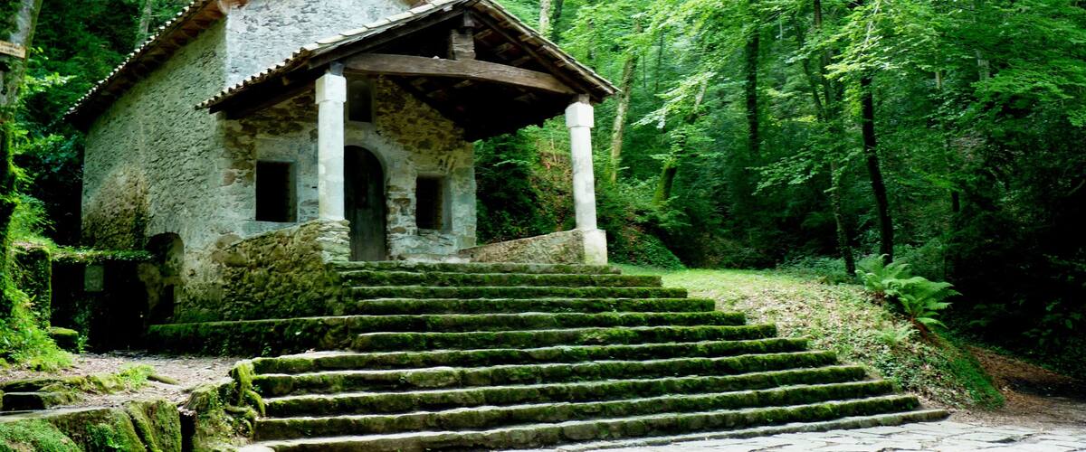 A stone chapel with a tiled roof, surrounded by a forested area with a stone staircase leading up to it.