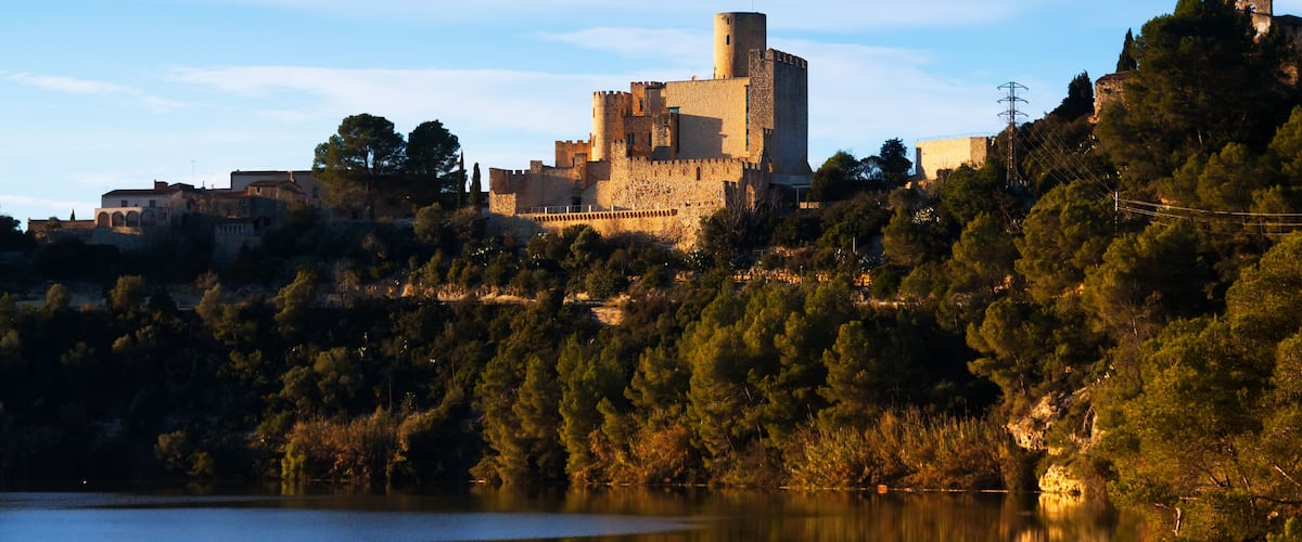 Un castillo de piedra histórico con una torre destacada, rodeado de un bosque denso y con vistas a un lago tranquilo.
