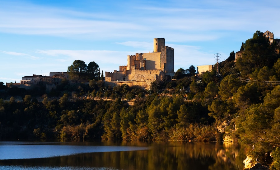 A historic stone castle with a prominent tower, surrounded by dense forest and overlooking a calm lake.