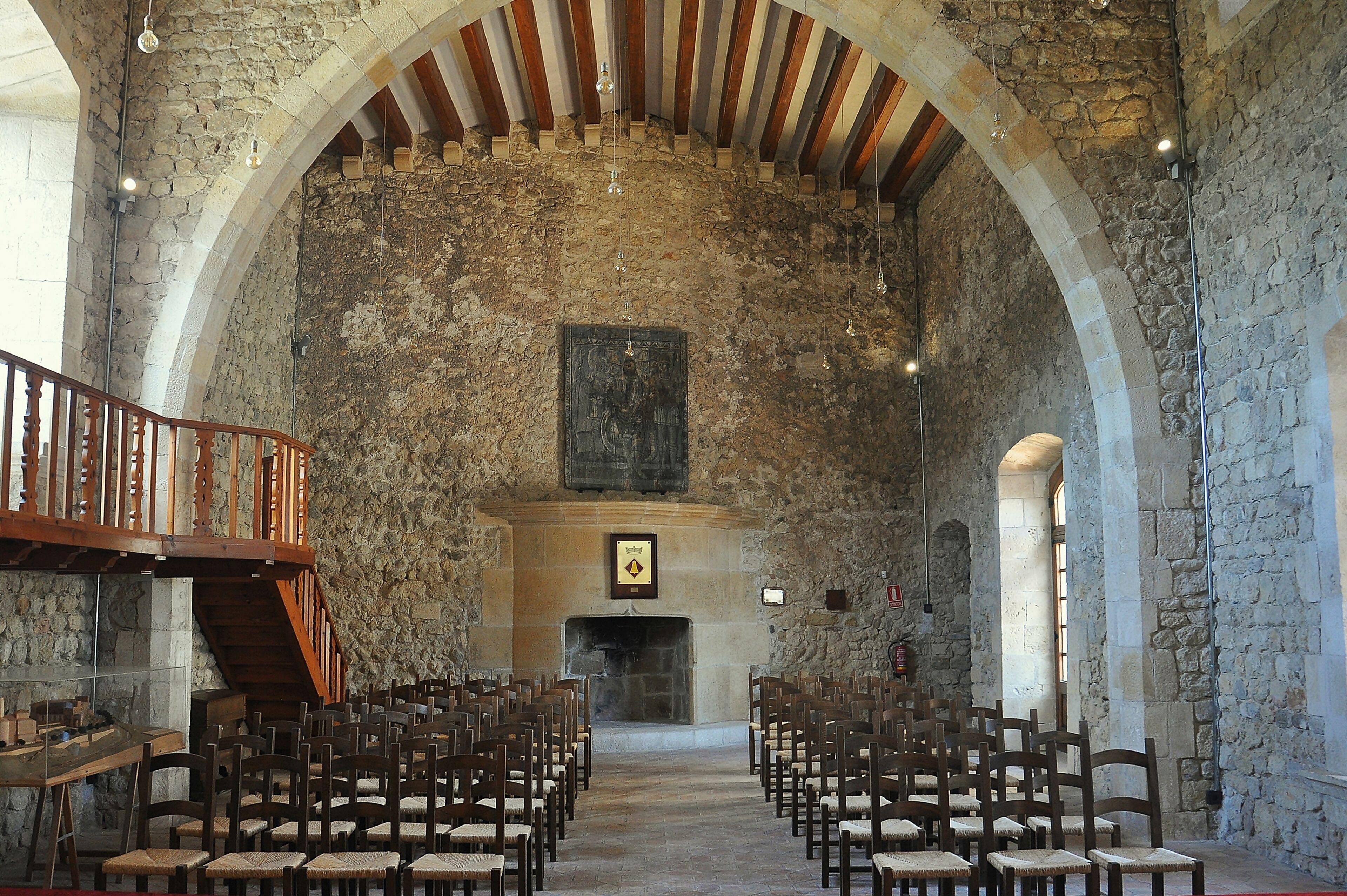 A stone-walled interior with wooden pews arranged in rows, a wooden staircase, and a fireplace.