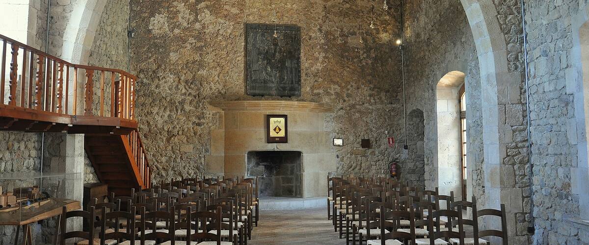 A stone-walled interior with wooden pews arranged in rows, a wooden staircase, and a fireplace.
