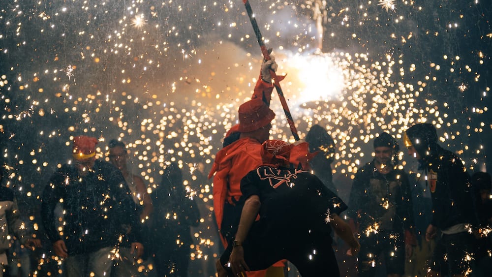 A person in a red jacket holding a stick amidst a crowd of people, with sparks flying around.