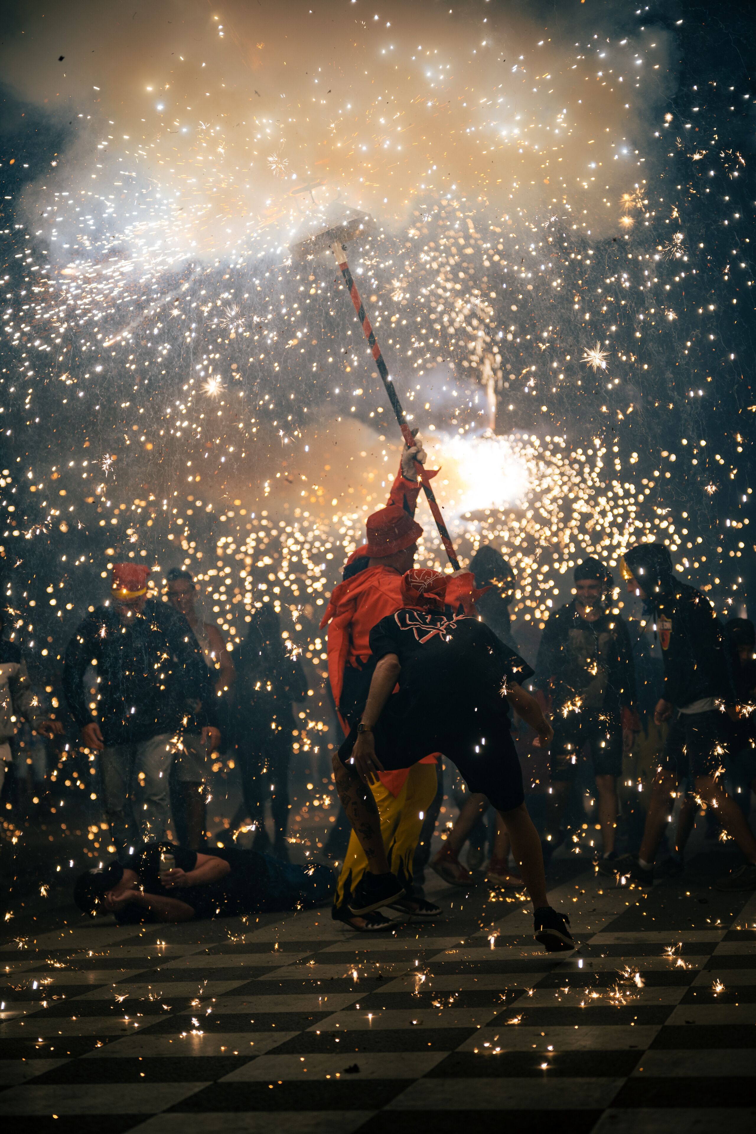 A person in a red jacket holding a stick amidst a crowd of people, with sparks flying around.