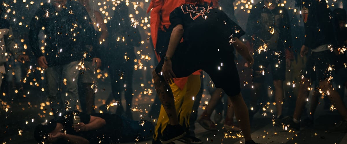 A person in a red jacket holding a stick amidst a crowd of people, with sparks flying around.