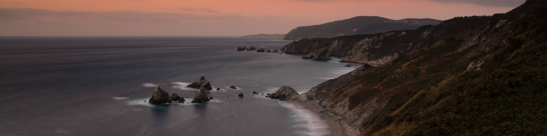 A coastal landscape with a curved shoreline, rocky outcrops, and a hilly terrain with sparse vegetation.