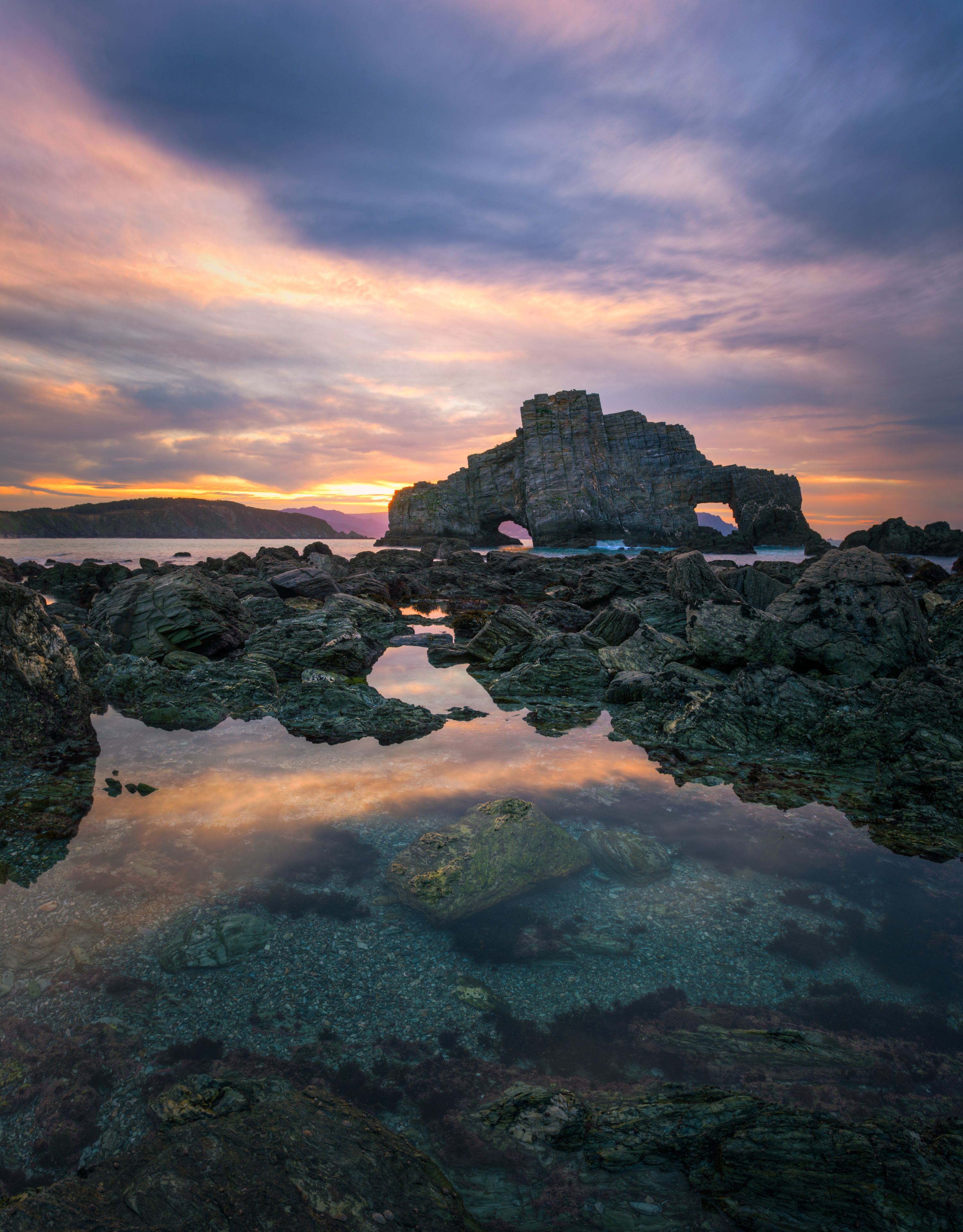 A rocky coastal landscape with a prominent rock formation in the center, surrounded by calm waters reflecting the sky.