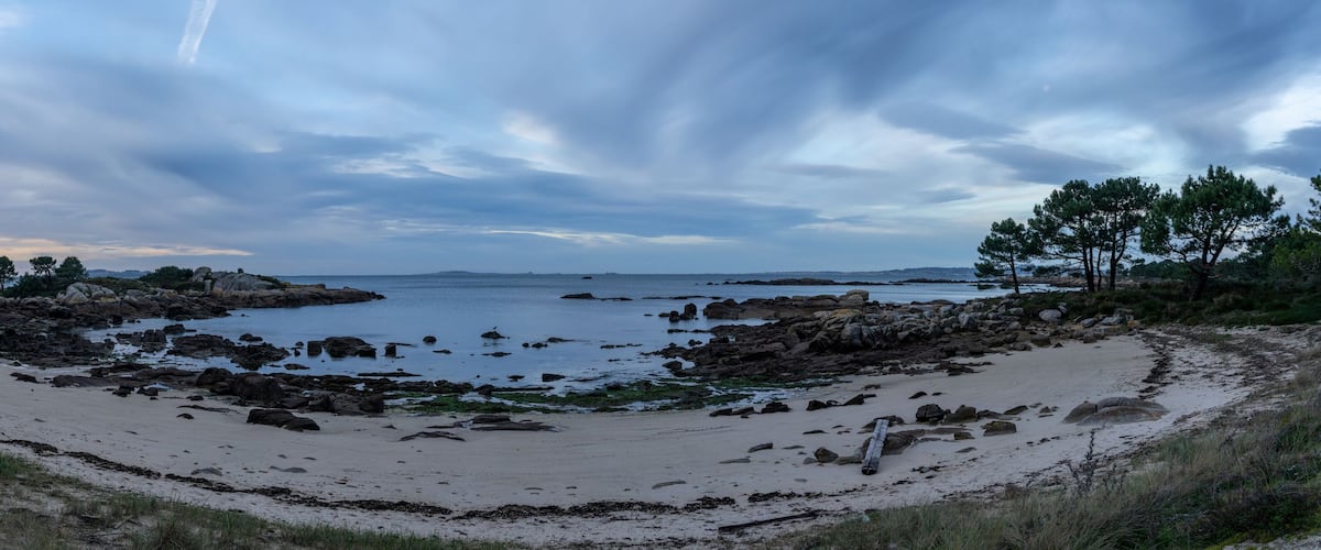 A coastal landscape with a sandy beach, rocky outcrops, and a calm sea under a cloudy sky.