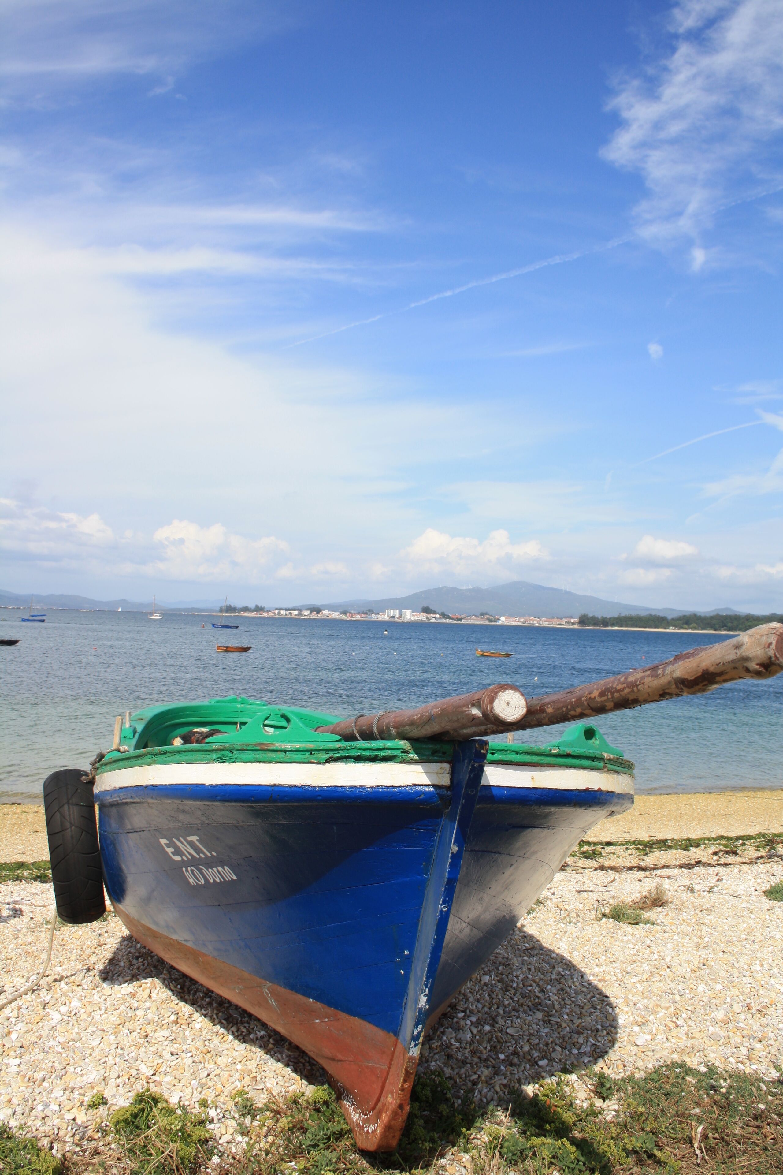 A blue and green boat with a rope tied to it, resting on a gravel beach with the sea and distant mountains in the background.