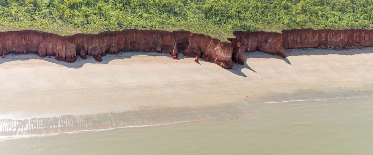 A coastal landscape with a sandy beach and a cliff with reddish-brown rock formations.