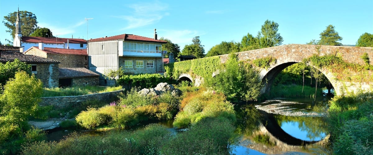A stone bridge over a river, surrounded by greenery and a building with a red roof.