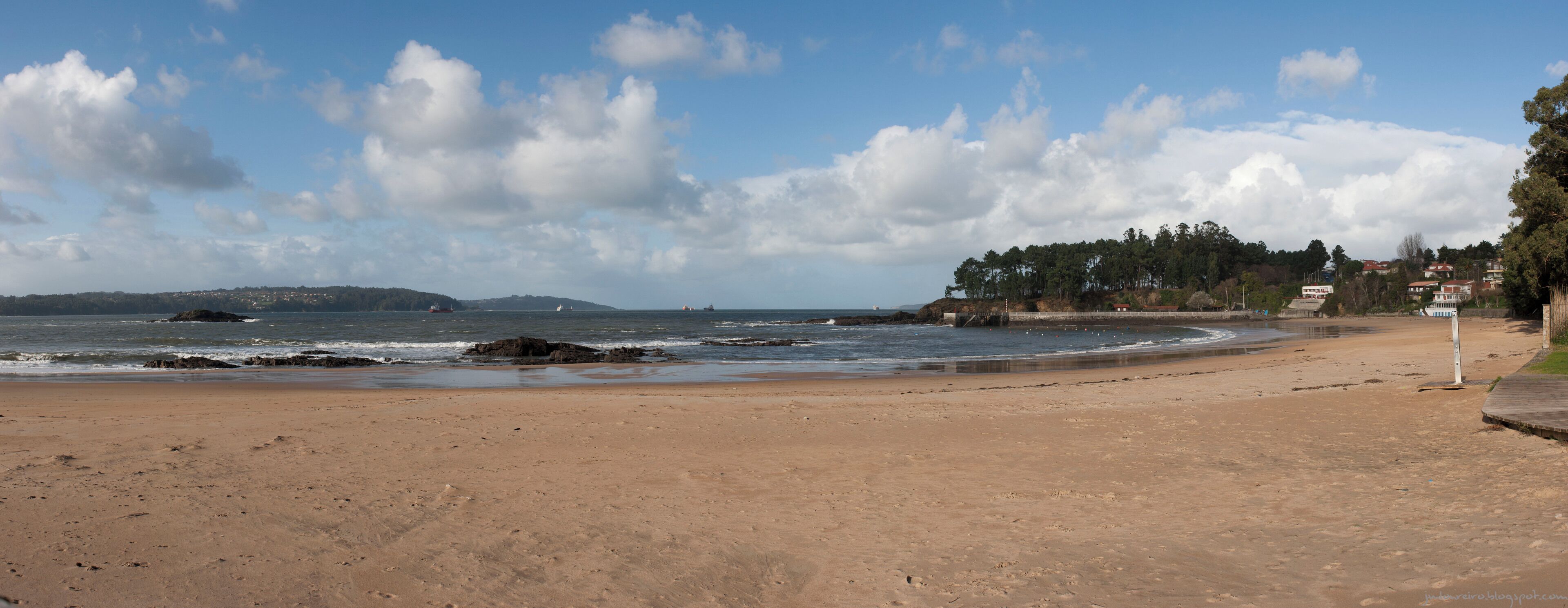 A sandy beach with waves, a rocky shoreline, and a forested hill in the background.