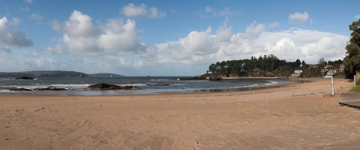 A sandy beach with waves, a rocky shoreline, and a forested hill in the background.