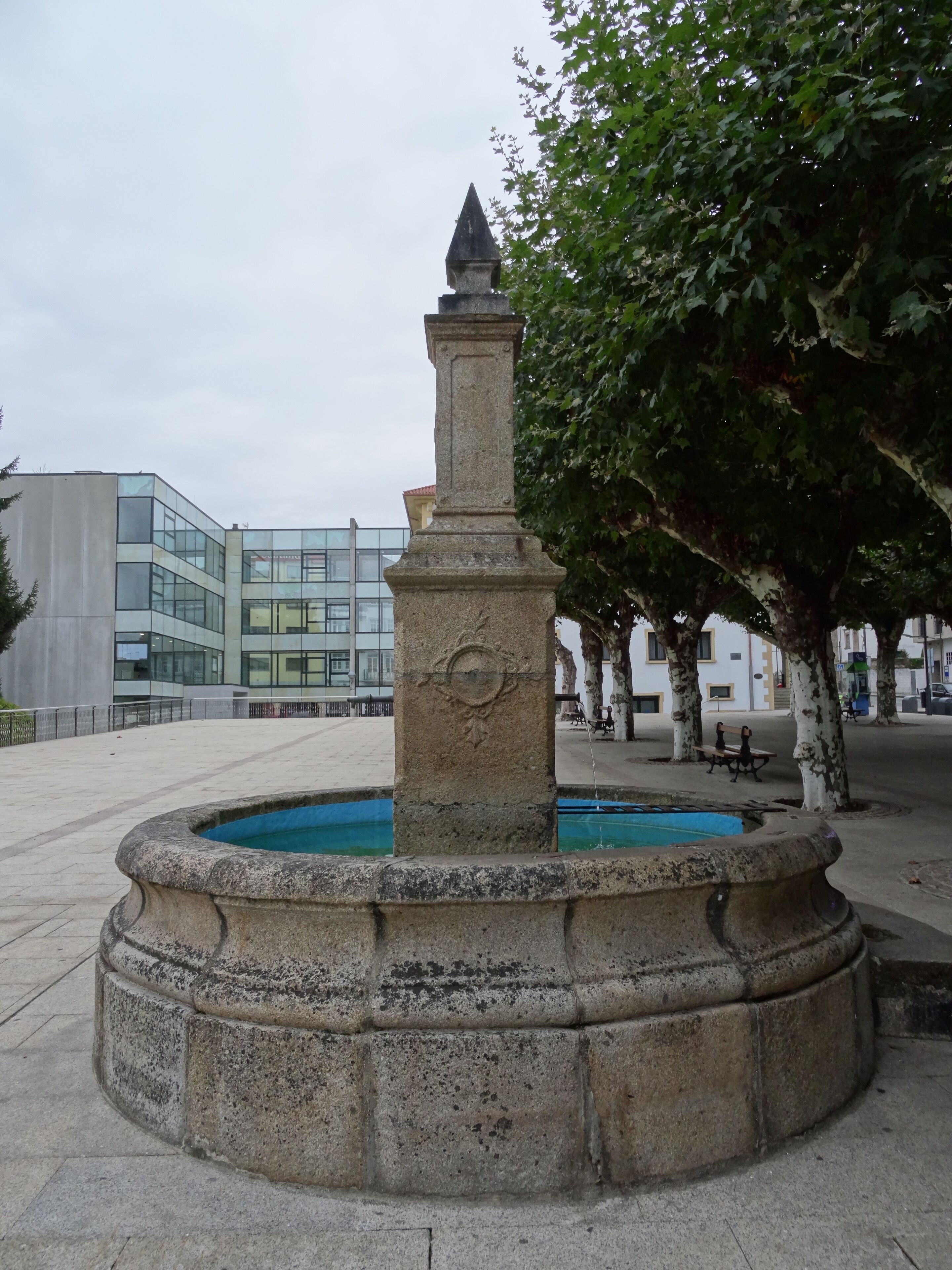 A stone fountain with a statue in the center, surrounded by a circular stone border. In the background, there is a modern building with glass windows and a tree with white bark.
