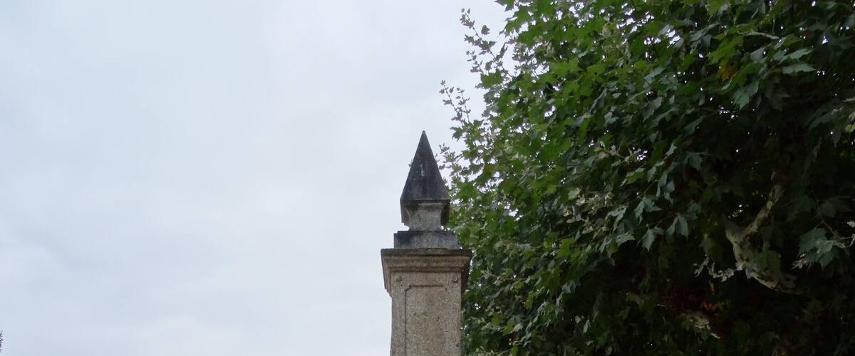 A stone fountain with a statue in the center, surrounded by a circular stone border. In the background, there is a modern building with glass windows and a tree with white bark.
