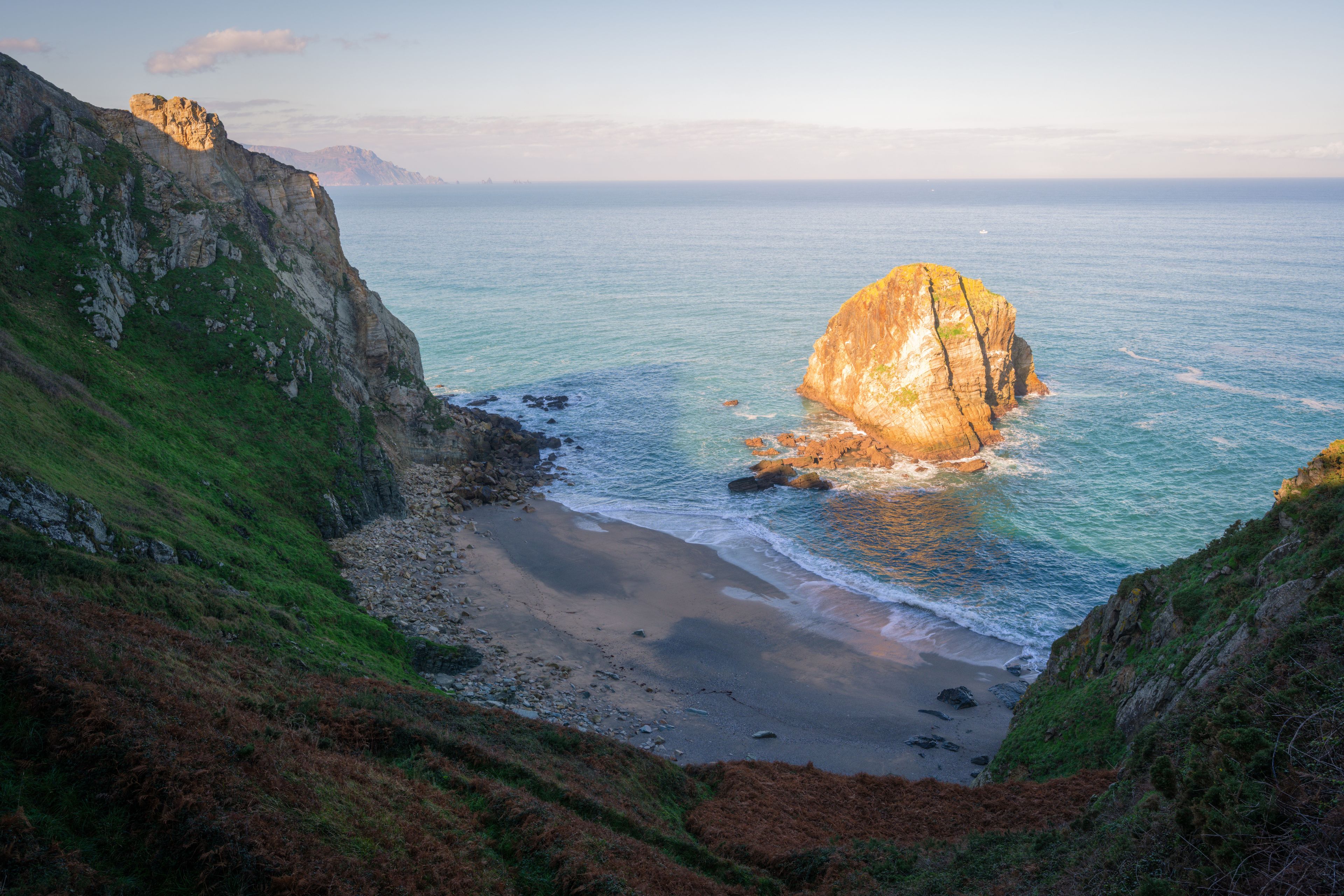Un paisaje costero con una formación rocosa destacada en el mar, rodeado de acantilados y una playa.
