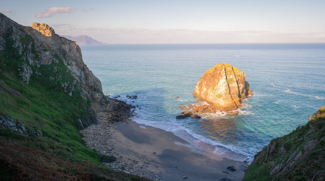 Un paisaje costero con una formación rocosa destacada en el mar, rodeado de acantilados y una playa.