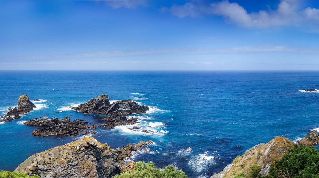 Un paisaje costero con acantilados rocosos, un mar azul cristalino y montañas lejanas bajo un cielo parcialmente nublado.