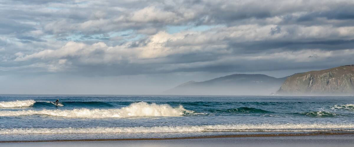 A surfer riding a wave near a rocky coastline under a cloudy sky.
