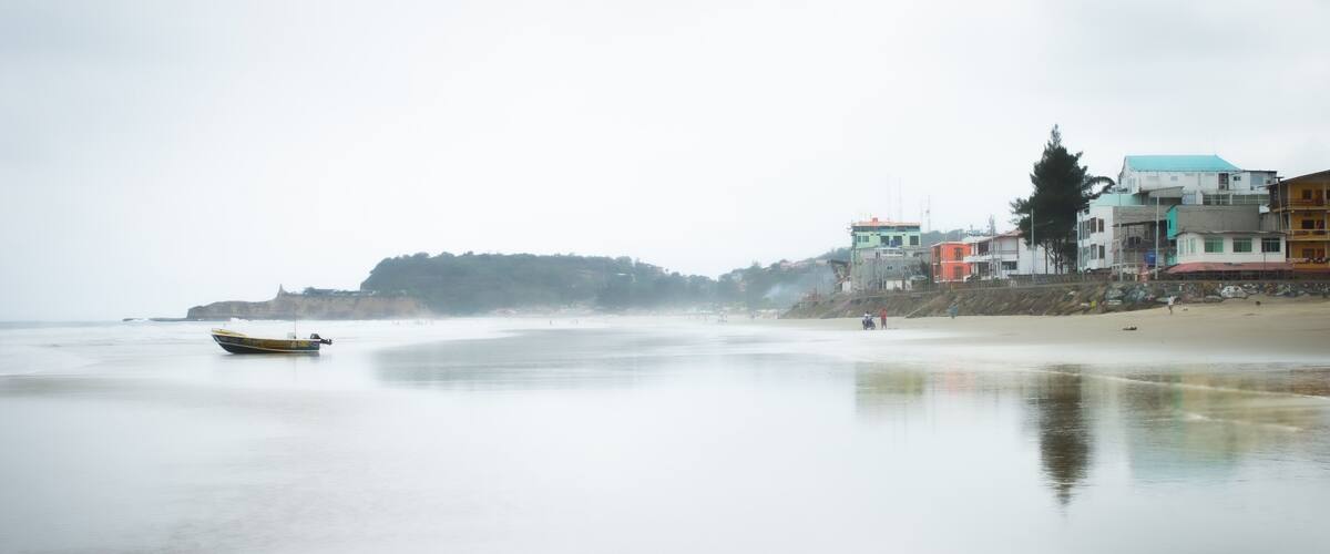A calm beach with a boat, buildings, and a hill in the background.