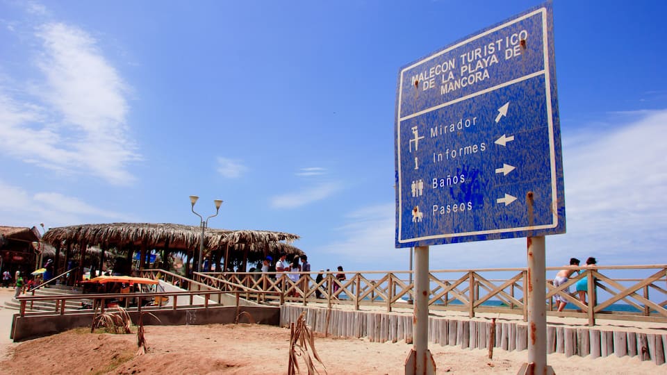 A beach with a sign indicating directions to Malecon, Playa de Mancora, and other attractions.