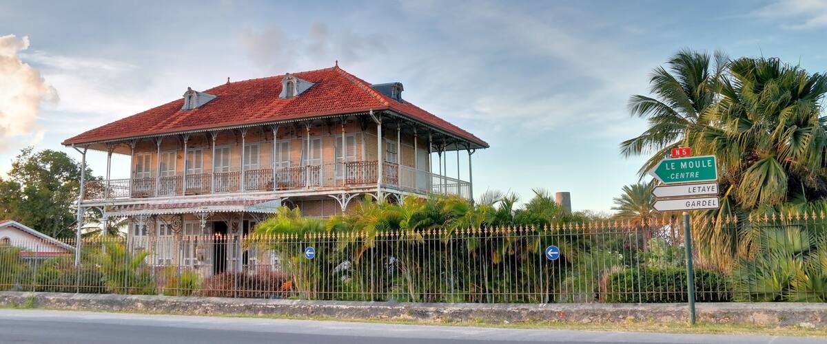 A historic building with a red-tiled roof and a balcony, surrounded by a metal fence and palm trees.