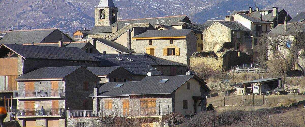 A village with stone buildings and a church with a bell tower, surrounded by snow-capped mountains.
