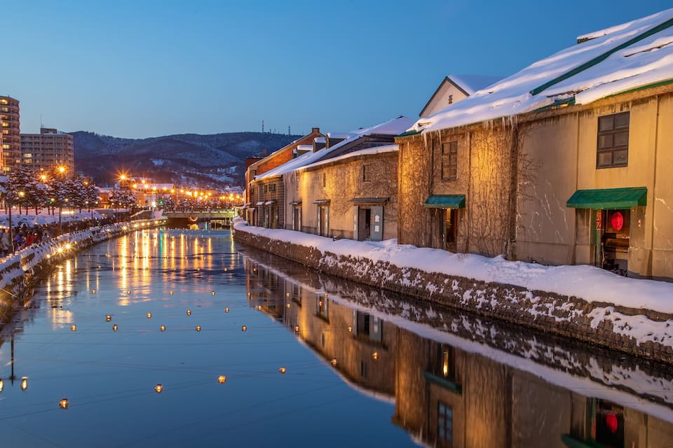 A snow-covered canal with reflections of lights, buildings, and a crowd of people.