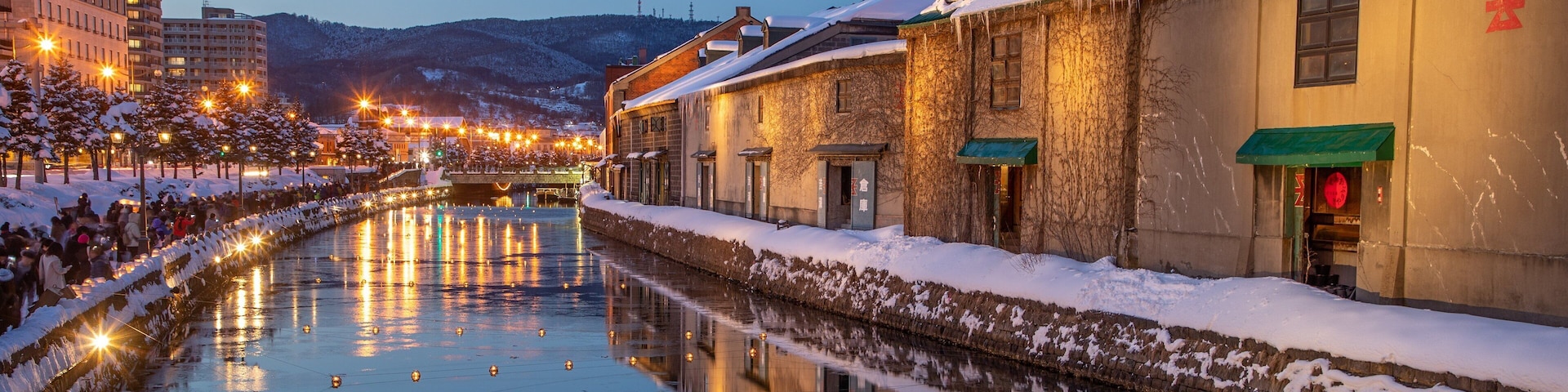 A snow-covered canal with reflections of lights, buildings, and a crowd of people.
