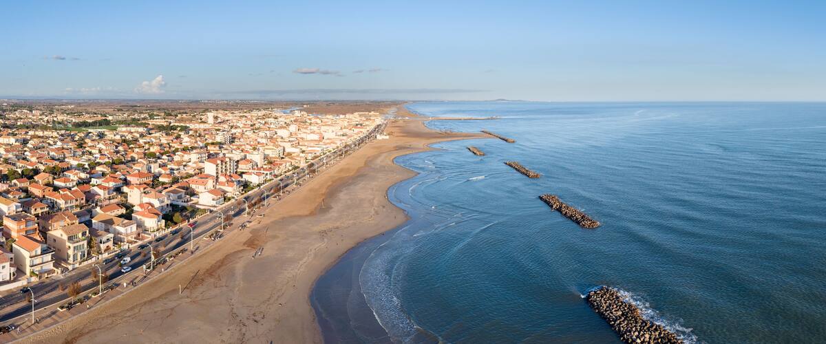 A coastal cityscape with a sandy beach, a clear blue sea, and a densely populated area with buildings.