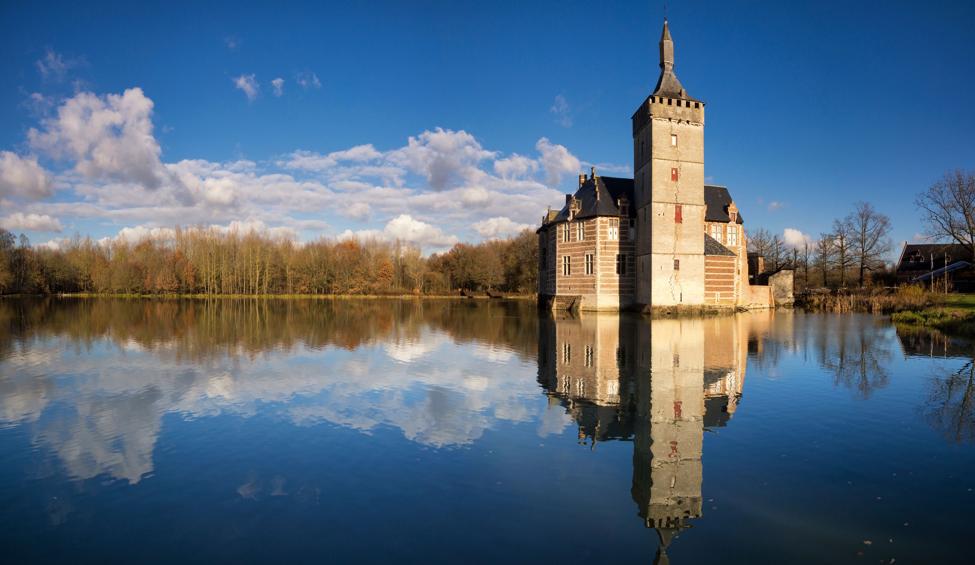Un castillo con una torre que se refleja en un lago tranquilo, rodeado de árboles y un cielo despejado.