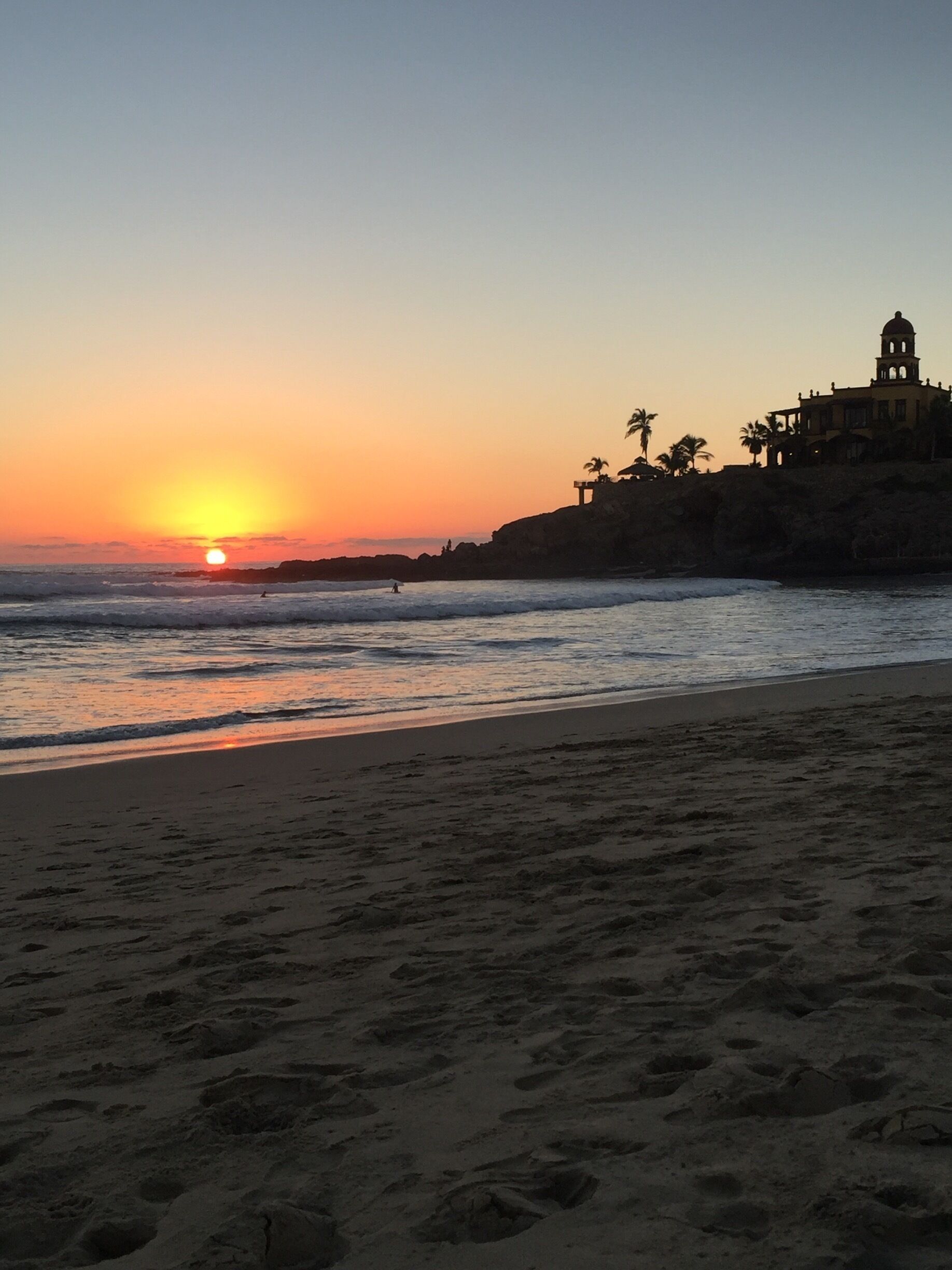 A sunset over a beach with footprints leading to the water, palm trees, and a building with a dome on a cliff.