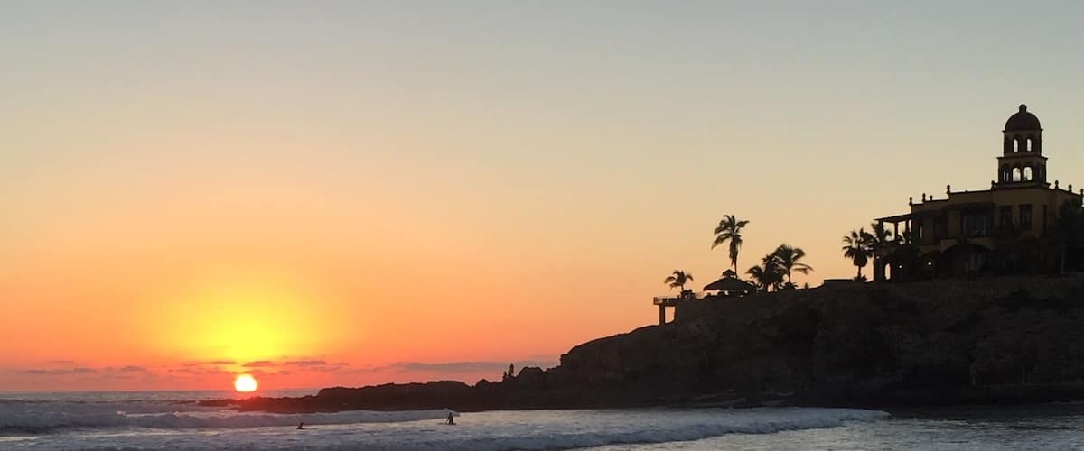 A sunset over a beach with footprints leading to the water, palm trees, and a building with a dome on a cliff.