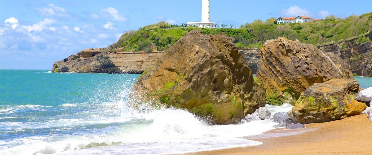 A lighthouse on a rocky cliff by the sea, with waves crashing against the rocks and a sandy beach in the foreground.
