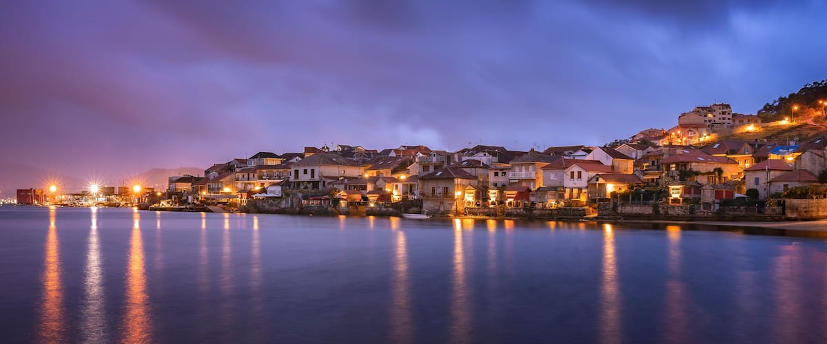 A coastal town at dusk with buildings reflecting on calm waters.