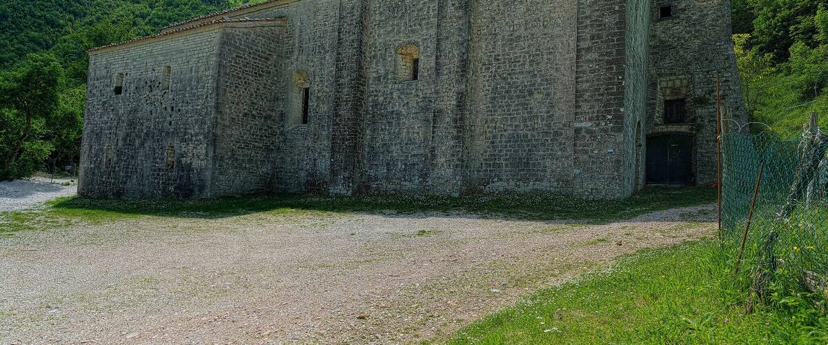 A large, weathered stone building with a flat roof, surrounded by lush green hills and a gravel path.