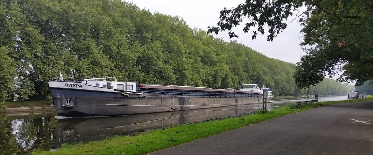A large cargo ship named 'RAYPA' docked along a riverbank with lush greenery and a paved road beside it.