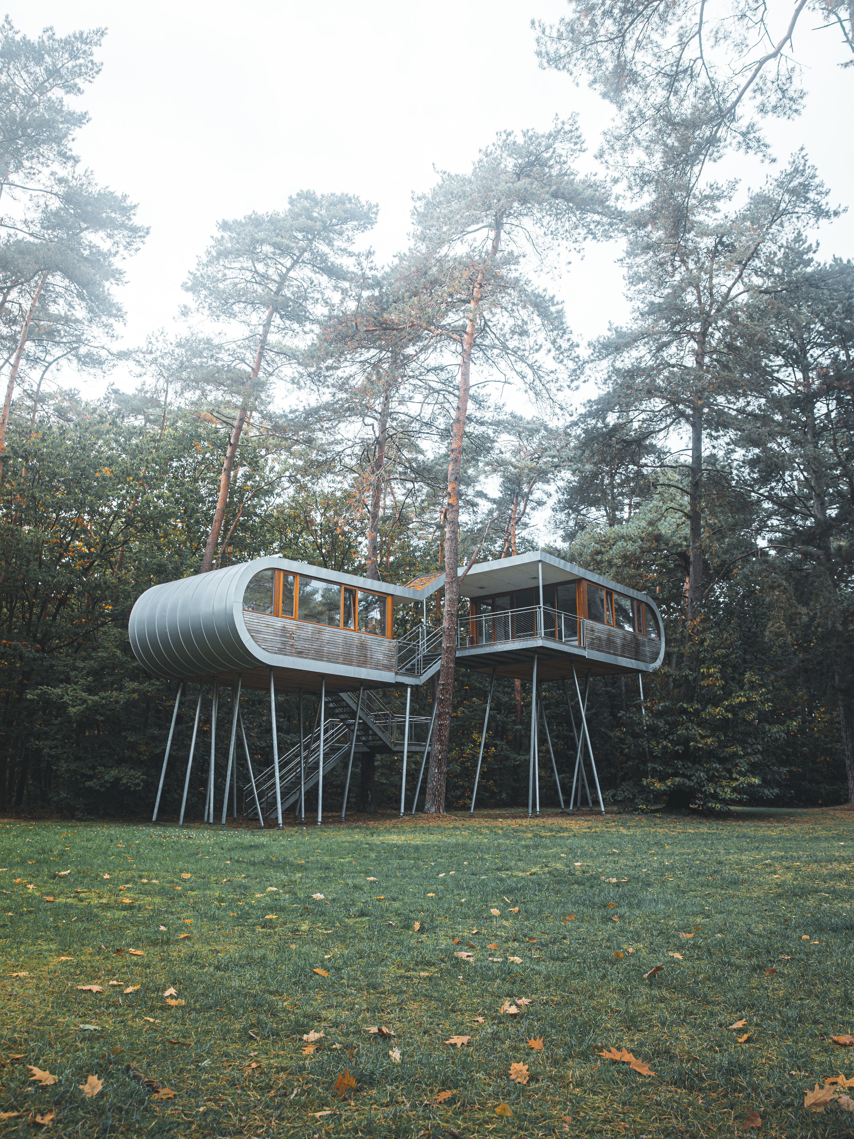 A modern treehouse structure with a cylindrical design, supported by metal stilts, surrounded by a forested area with tall trees and a grassy field.