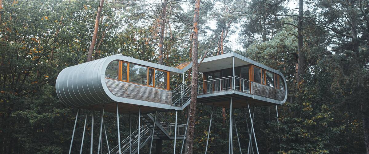 A modern treehouse structure with a cylindrical design, supported by metal stilts, surrounded by a forested area with tall trees and a grassy field.