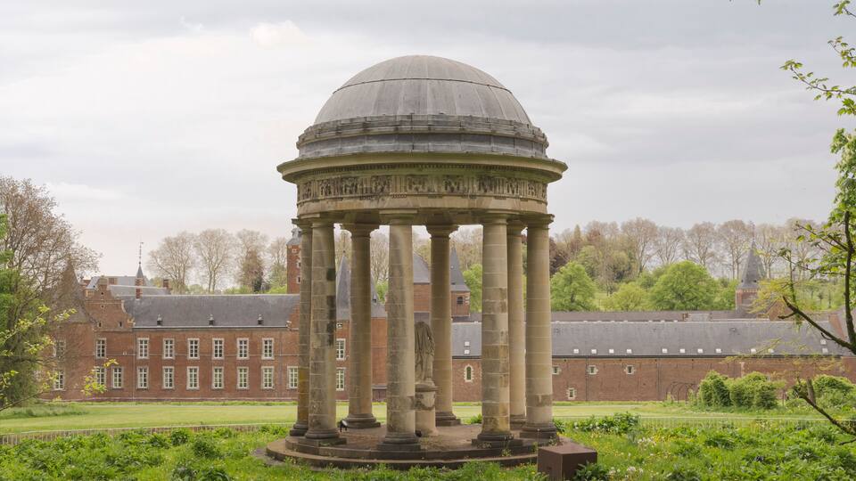 A classical pavilion with a dome and columns, surrounded by lush greenery and a large brick building in the background.
